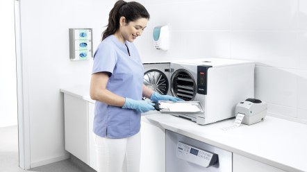 Eine medizinische Fachangestellt steht vor einer Aufbereitungszeile, wo ein Reinigungs- und Desinfektionsgerät sowie ein Sterilisator stehen.